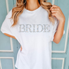 A women's graphic t-shirt with short sleeves, featuring the word "Bride" in a floral design. This soft and comfortable Comfort Colors short sleeve tee is ideal for celebrating bridal engagements. Available in white, in sizes from Small to 2XL.